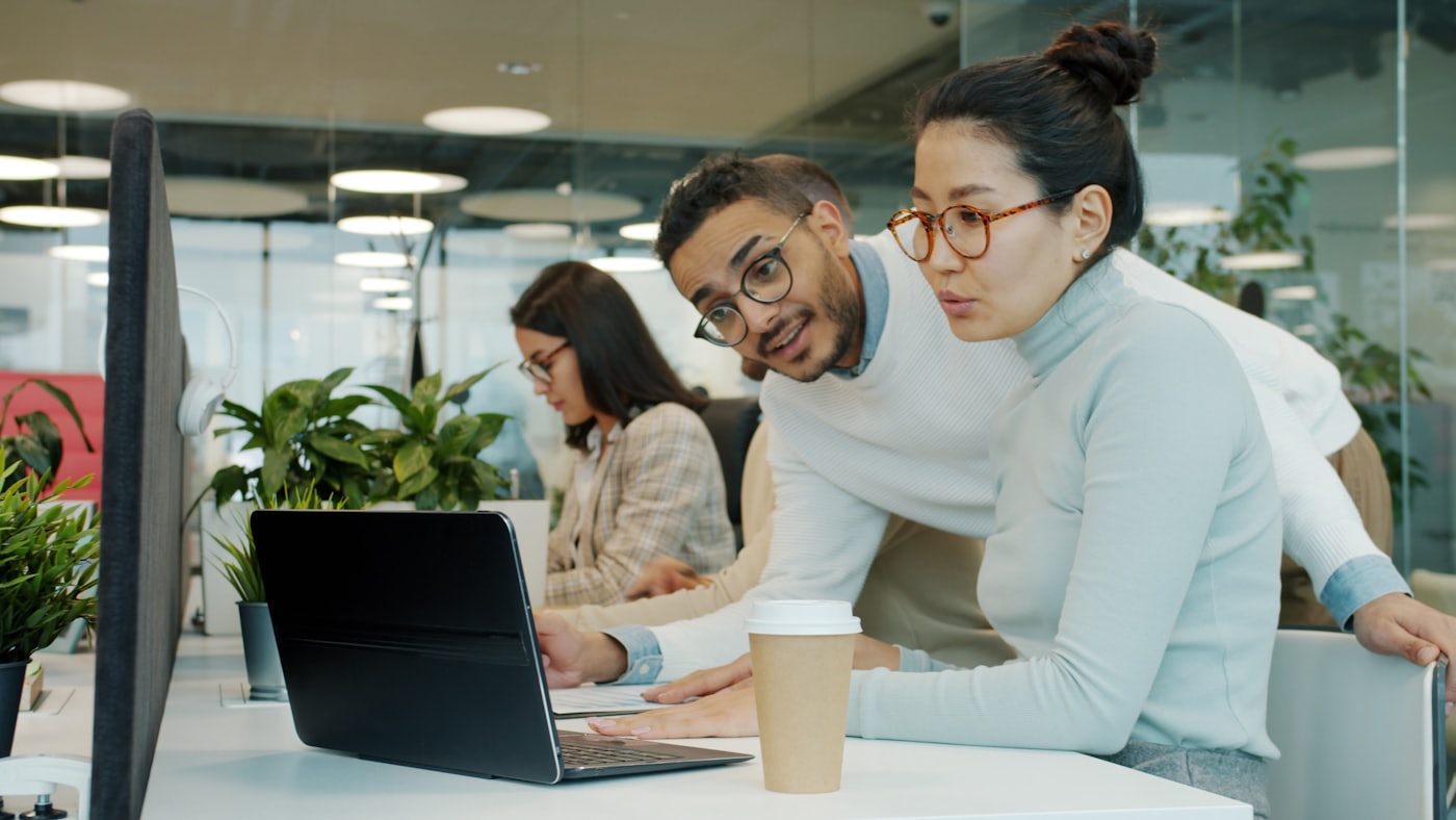 Two colleagues leaning over one laptop, talking through a problem.